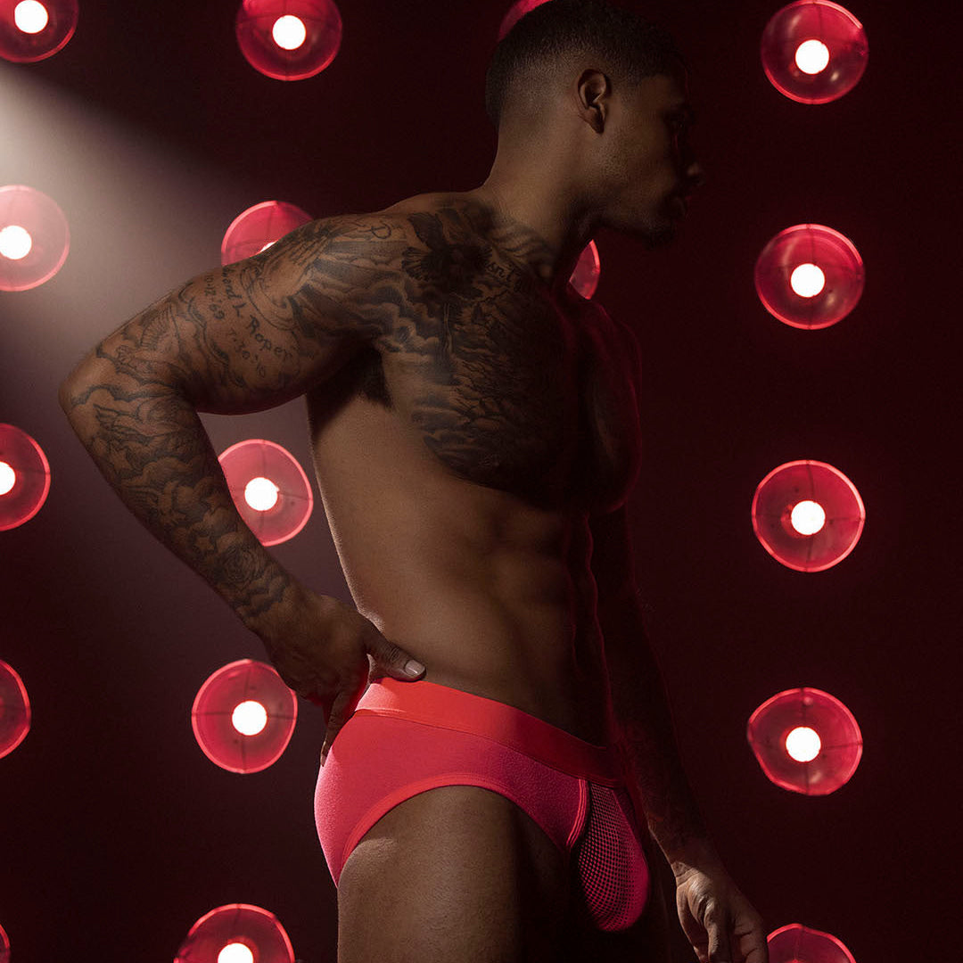 A shirtless tattooed man in bright red underwear stands in profile against a dark background with circular red lights.