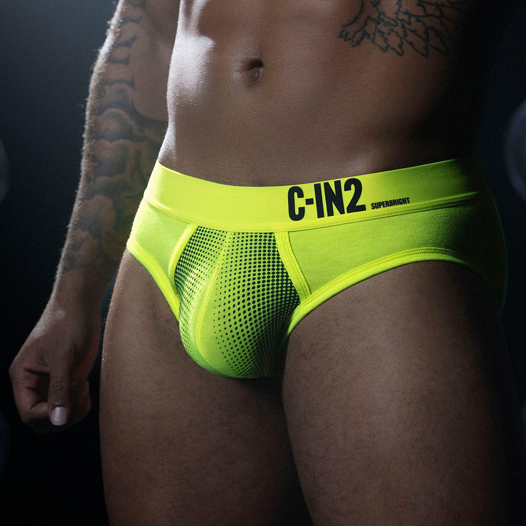 A close-up of a model's lower torso wearing bright neon yellow-green mesh briefs with a wide waistband and black 'C-IN2' branding, photographed against a dark studio background.