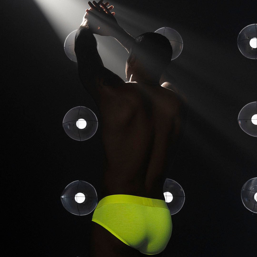 A model shown from behind wearing neon yellow-green briefs, standing in a dark studio with dramatic spotlight beams and circular light fixtures on a black background.