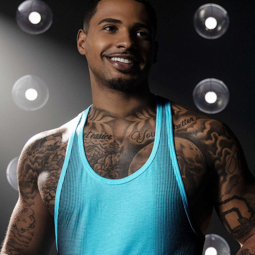 Man wearing a blue tank top with tattoos, standing against a dark background with light bulbs.