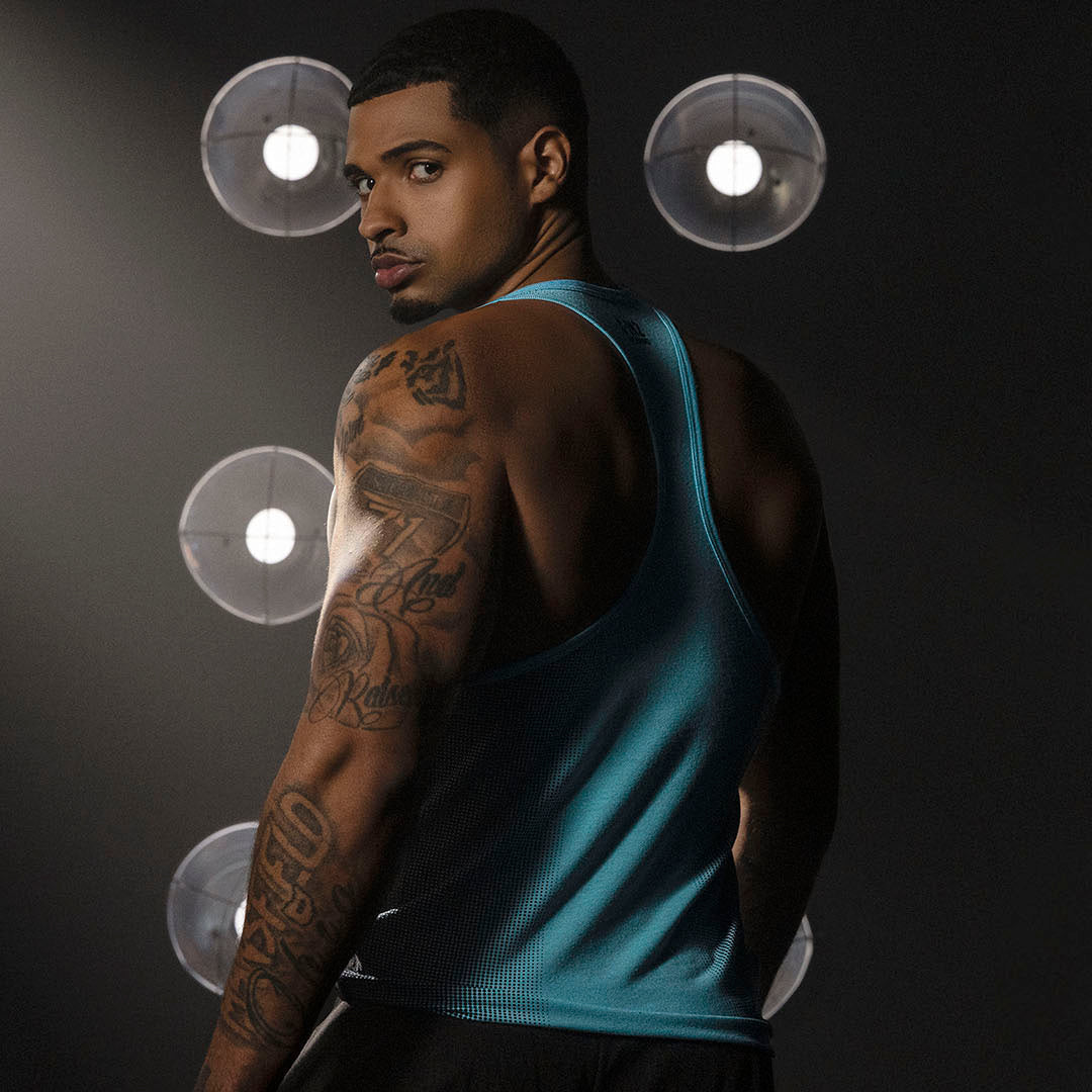 Man wearing a blue tank top with tattoos on his arms, standing against a dark background with light fixtures.