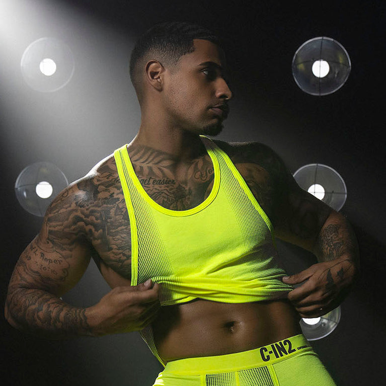 A tattooed male model wearing a bright neon yellow-green ribbed tank top and matching briefs with 'C-IN2' on the waistband, posed against a dark background with spotlight effects and circular light reflections.