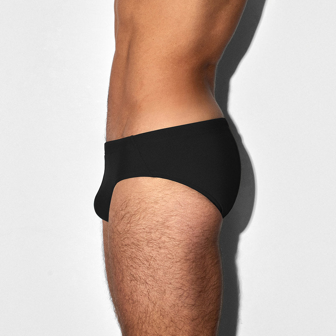 A side profile view of a man's lower torso and upper thigh wearing a pair of black swim briefs against a light gray background.