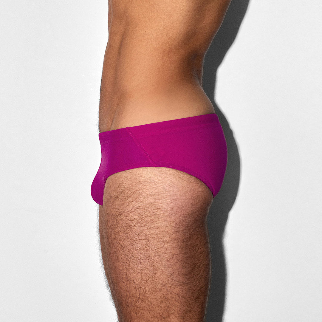 A side profile view of a man's lower torso and upper thigh wearing bright raspberry-pink swim briefs against a light gray background.