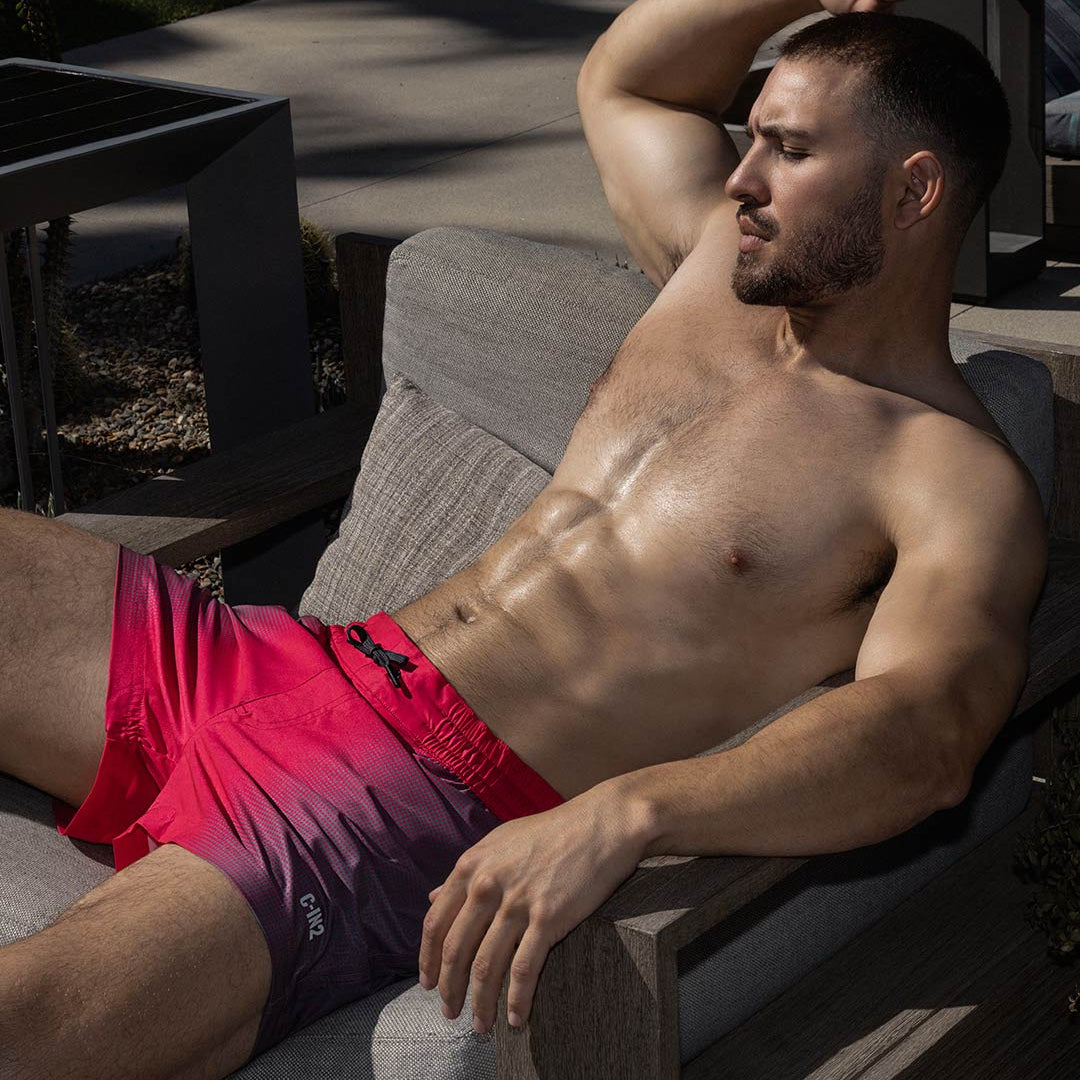 Man in red swim shorts with 'C-IN2' logo lying on a wooden bench outdoors