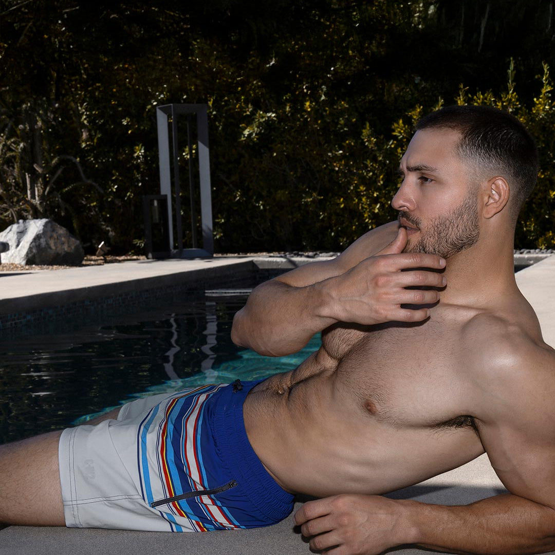 Man in blue C-IN2 swim trunks sitting by a pool with trees in the background