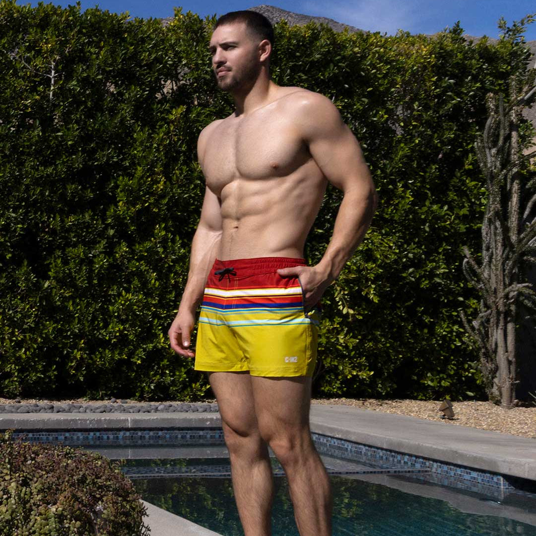 Man wearing colorful swim shorts with C-IN2 branding standing by a pool with greenery in the background