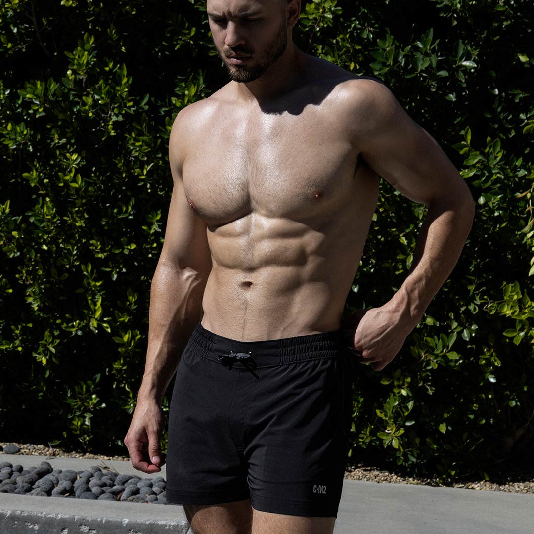 Man with a muscular build wearing black swim shorts with 'C-IN2' logo standing against a green hedge.