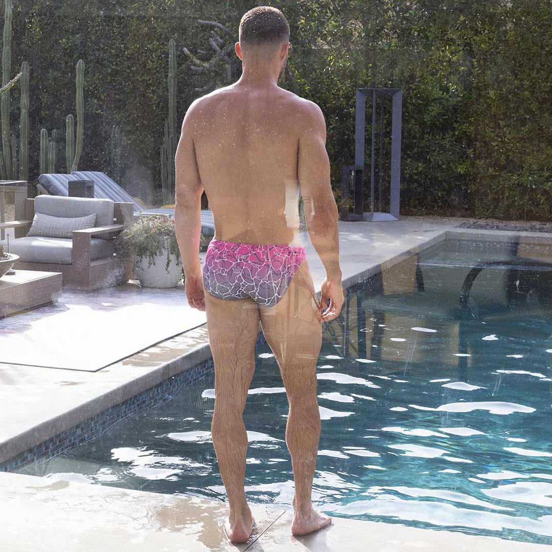 Man standing by a pool wearing pink swim briefs with a pattern.