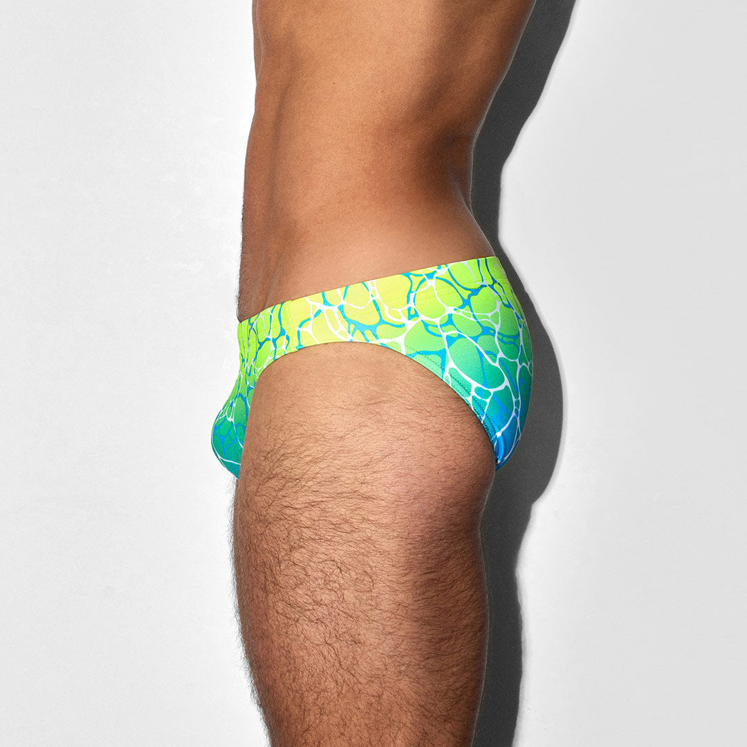A side profile view of a man wearing swim briefs with a vibrant green, yellow, and blue water-ripple pattern against a light gray background.