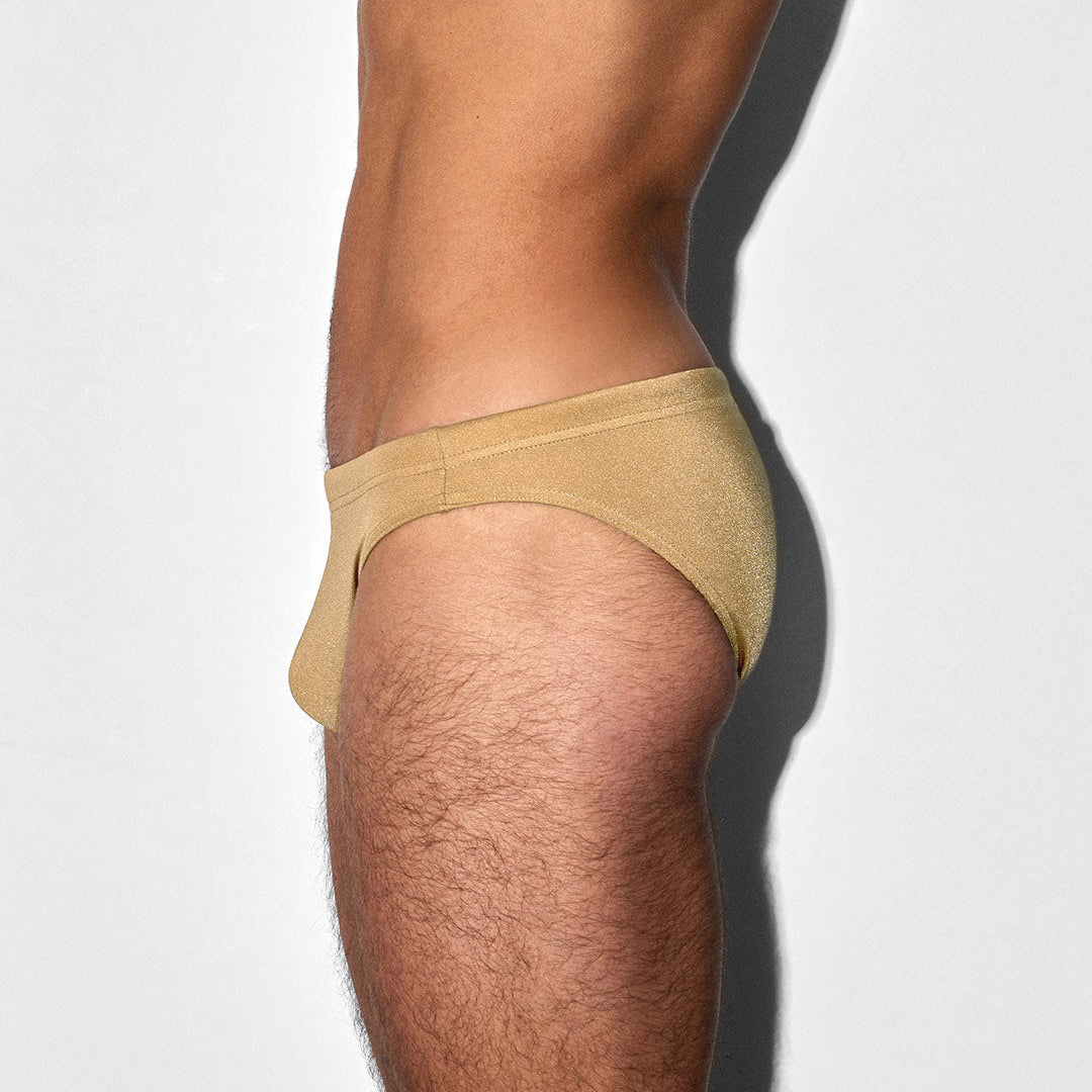 A side profile view of a man's lower torso and upper thigh wearing a pair of low-rise, light tan/beige briefs against a plain white background.