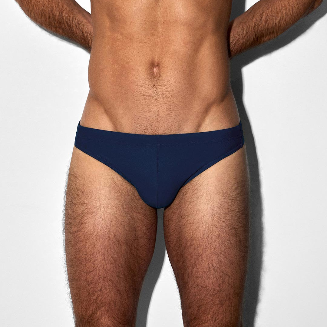A close-up of a male model's torso wearing a pair of navy blue tanga swim briefs against a plain white background.