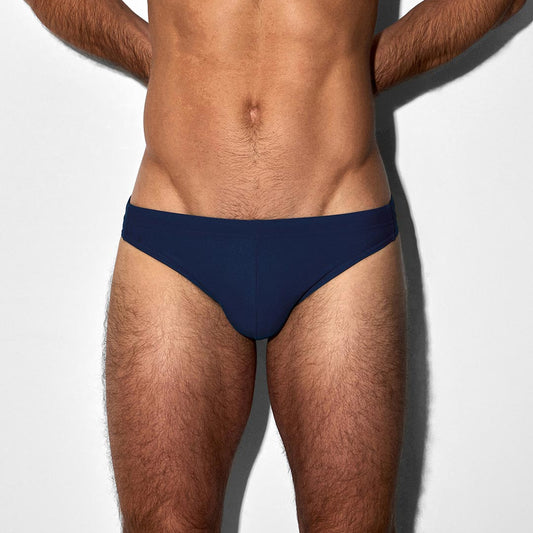 A close-up of a male model's torso wearing a pair of navy blue tanga swim briefs against a plain white background.