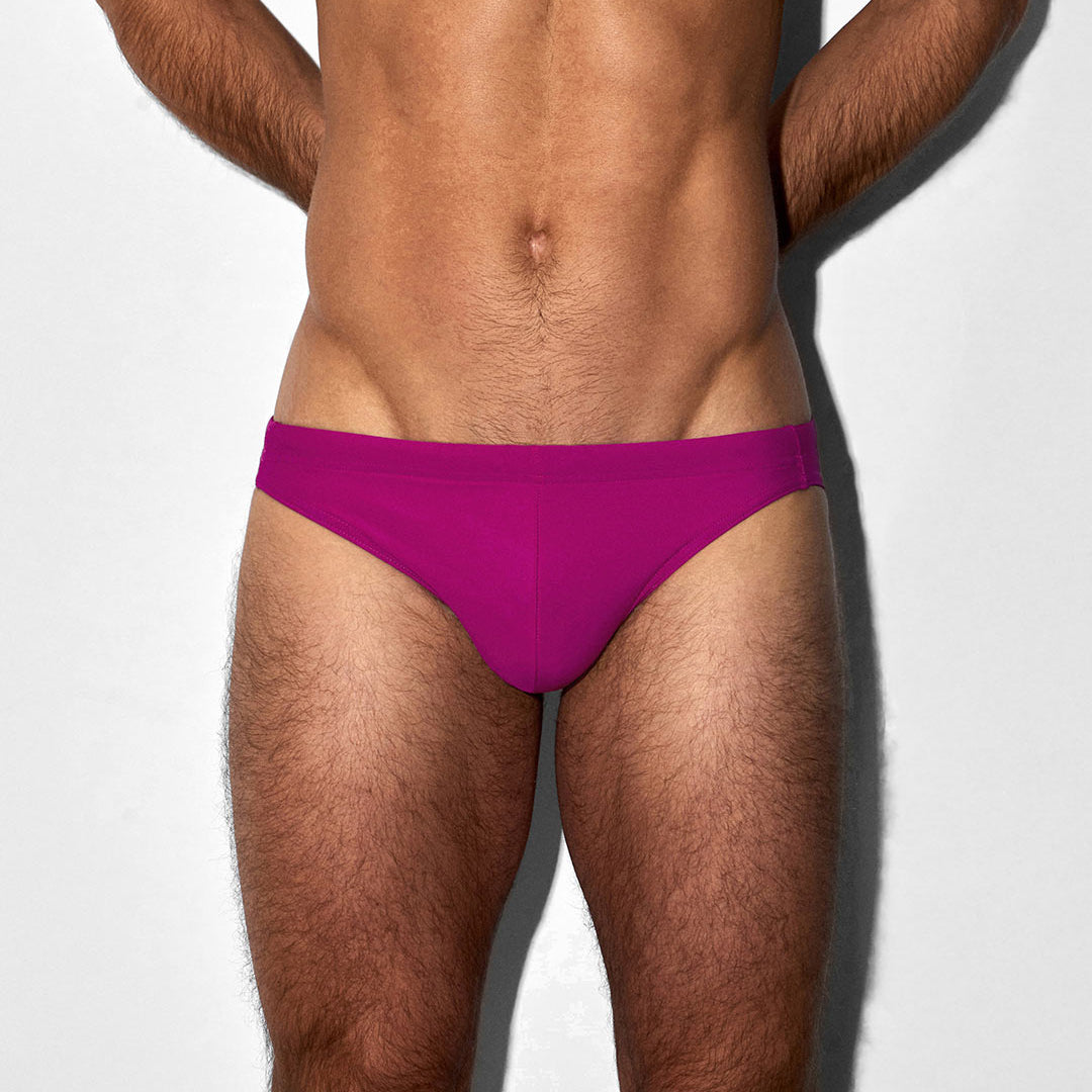 A close-up of a male model's lower torso wearing bright raspberry-pink swim briefs against a plain light gray background.