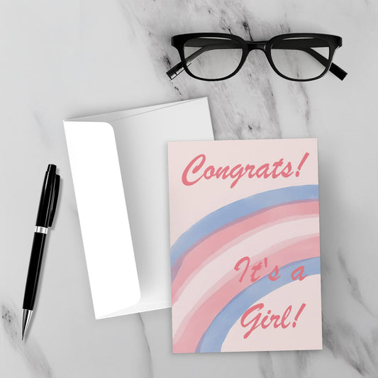 Greeting card with trans rainbow design and 'Congrats! It's a Girl!' text on a marble surface with glasses and pen.