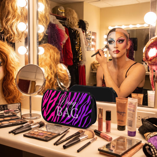 A vanity table in a dressing room with a black makeup bag featuring a bright pink and purple zebra print and the words 'Drag BAG' in white script. A drag queen with dramatic eye makeup applies makeup in front of a lighted mirror, with wigs, clothing, and additional cosmetics visible around her.