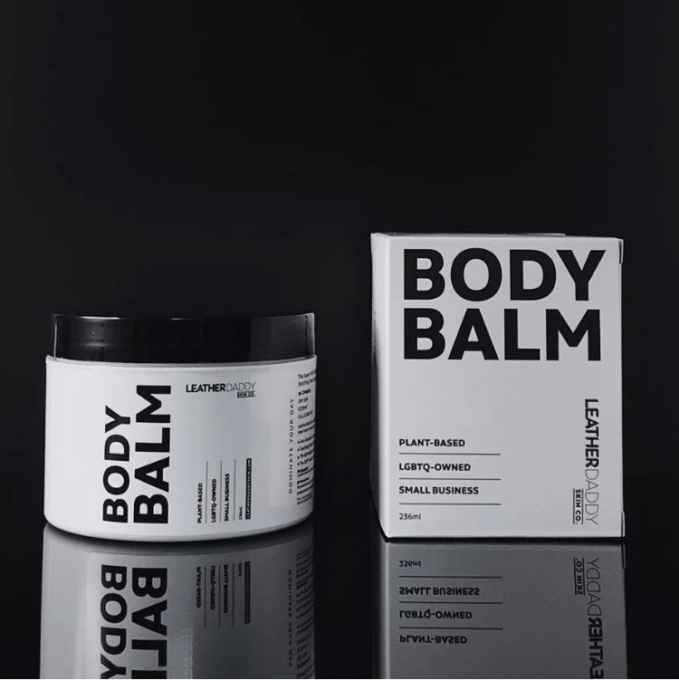 White jar with black lid and black lettering of body balm and leather daddy branding on a black reflective surface