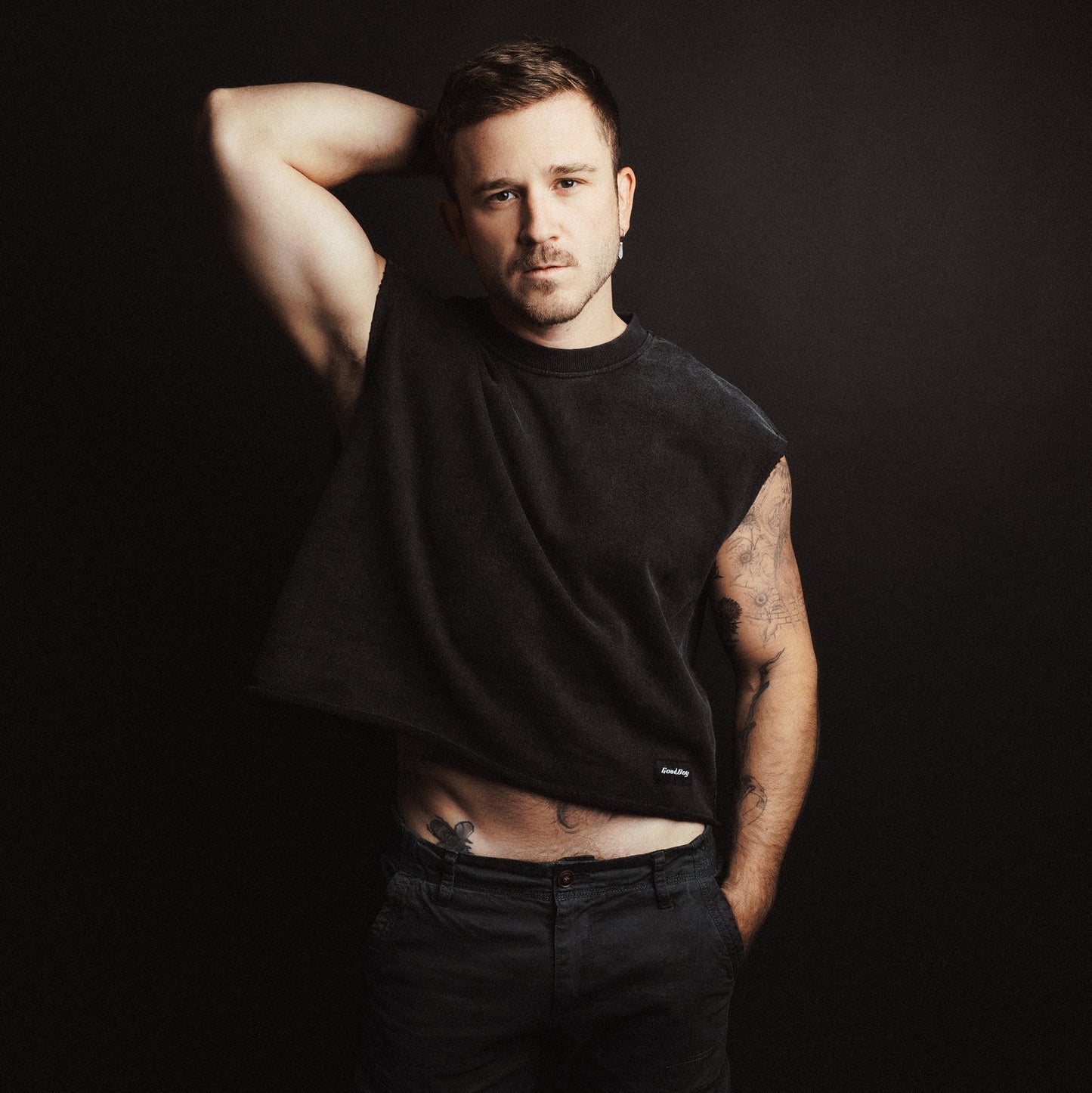 Man wearing a black cropped t-shirt with 'Good Boy' branding and pants against a dark background