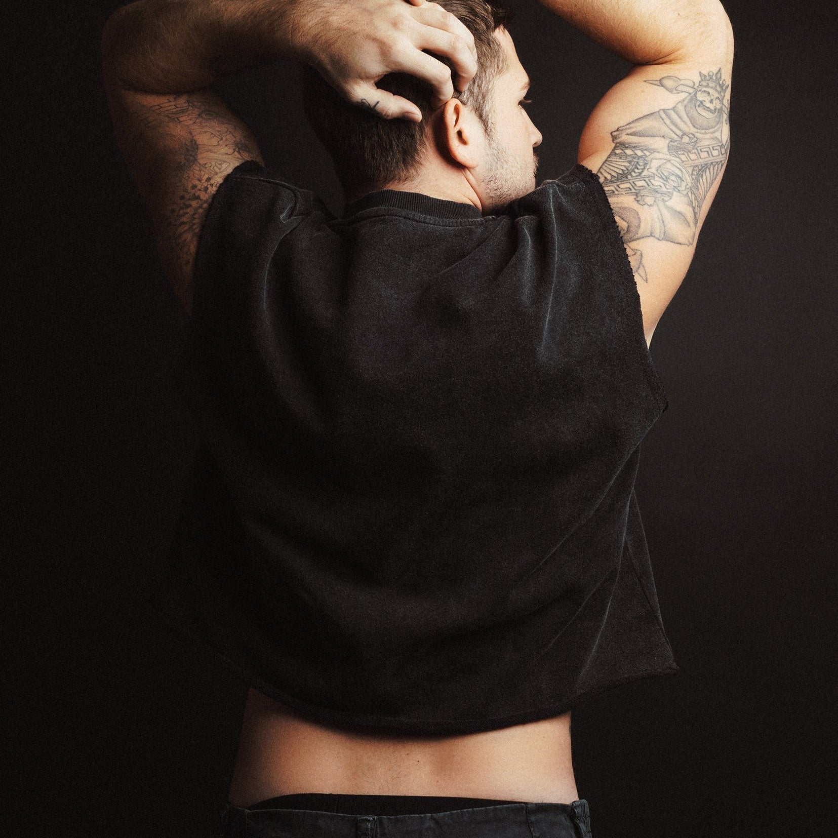 Back of man with tattoos on arms and back wearing a black cropped t-shirt against a dark background