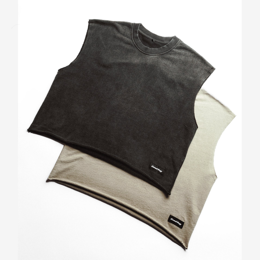 Two sleeveless cropped t-shirts laid flat on a white background, one in washed black and one in washed beige. Both shirts feature a crew neck and a small black 'Good Boy' brand label near the bottom hem.