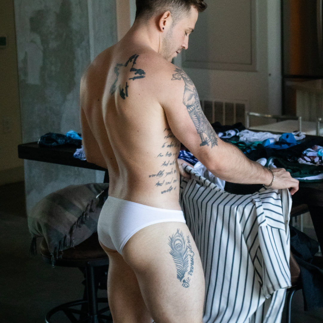 Man with tattoos wearing white underwear in a room with striped pants and clothing on a table.