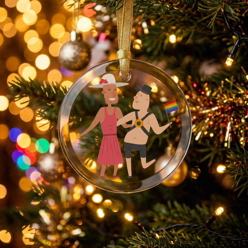 glass holiday ornament hanging on a christmas tree with lights.  The ornament is of two men dancing, one wearing a hat dress and the other wearing a leather harness and carrying an lgbtq rainbow pride flag