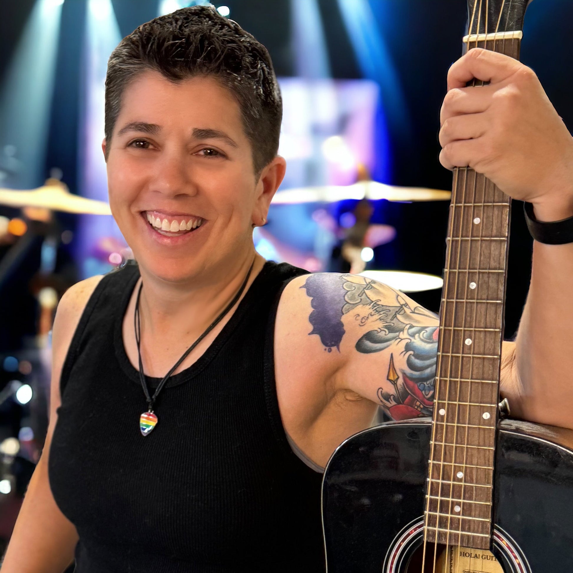 Person wearing an lgbtq pride guitar pick necklace holding a guitar with a blurred stage background
