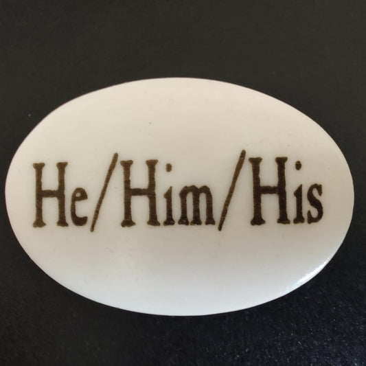 An oval white button or badge featuring the text 'He/Him/His' in dark brown serif lettering, set against a dark textured background.