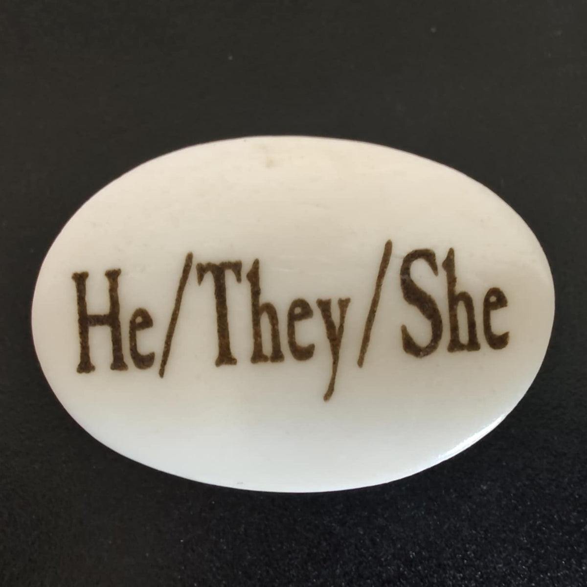An oval white bone charm with the words 'He/They/She' engraved in a dark brown serif font, shown on a black background.