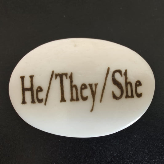 An oval white bone charm with the words 'He/They/She' engraved in a dark brown serif font, shown on a black background.