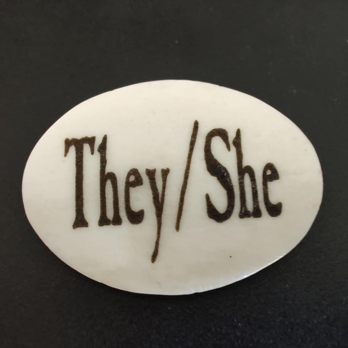 An oval lapel pin featuring the words 'They / She' in dark brown gothic-style lettering, set against a dark black background.