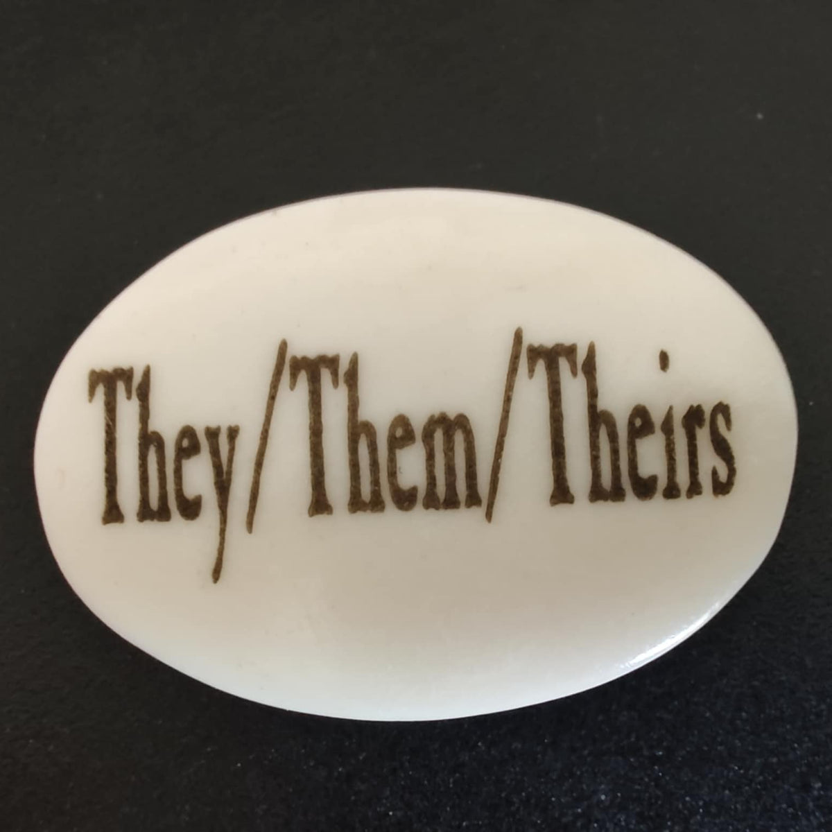 An oval white lapel pin with brown text reading 'They/Them/Theirs' on a dark background.