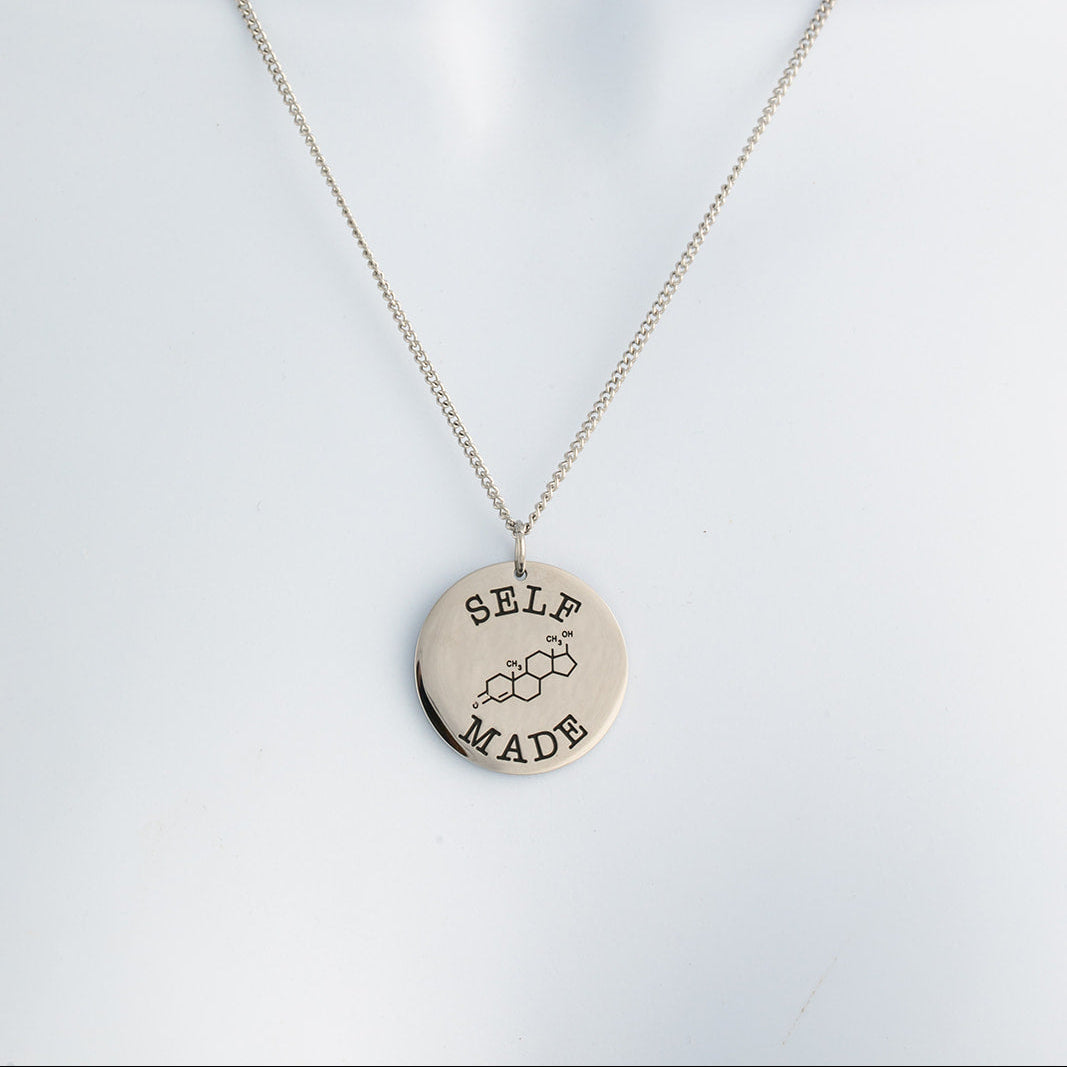 Polished silver necklace with a round pendant with text that says 'self made' and the chemical symbol for testosterone on a light gray background