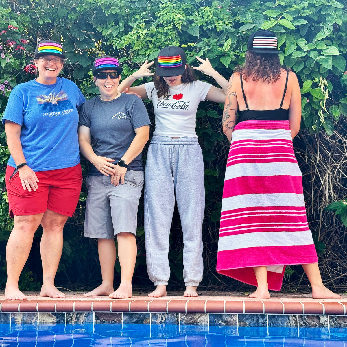 Four people wearing embroidered pride hats, standing by a pool with lush greenery in the background