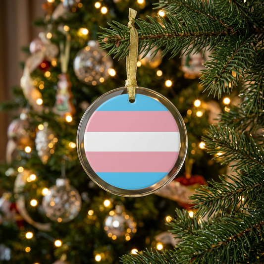 Round clear glass holiday ornament with the transgender pride flag colors in the center hung from a Christmas tree with lights