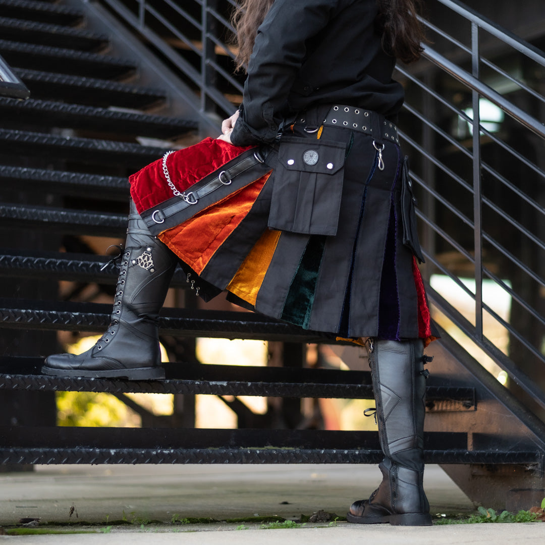 A person wearing a colorful rag-kilt skirt with red, orange, yellow, green, and blue panels, black leather utility belt and pouches, and tall black lace-up boots, standing on black metal stairs.