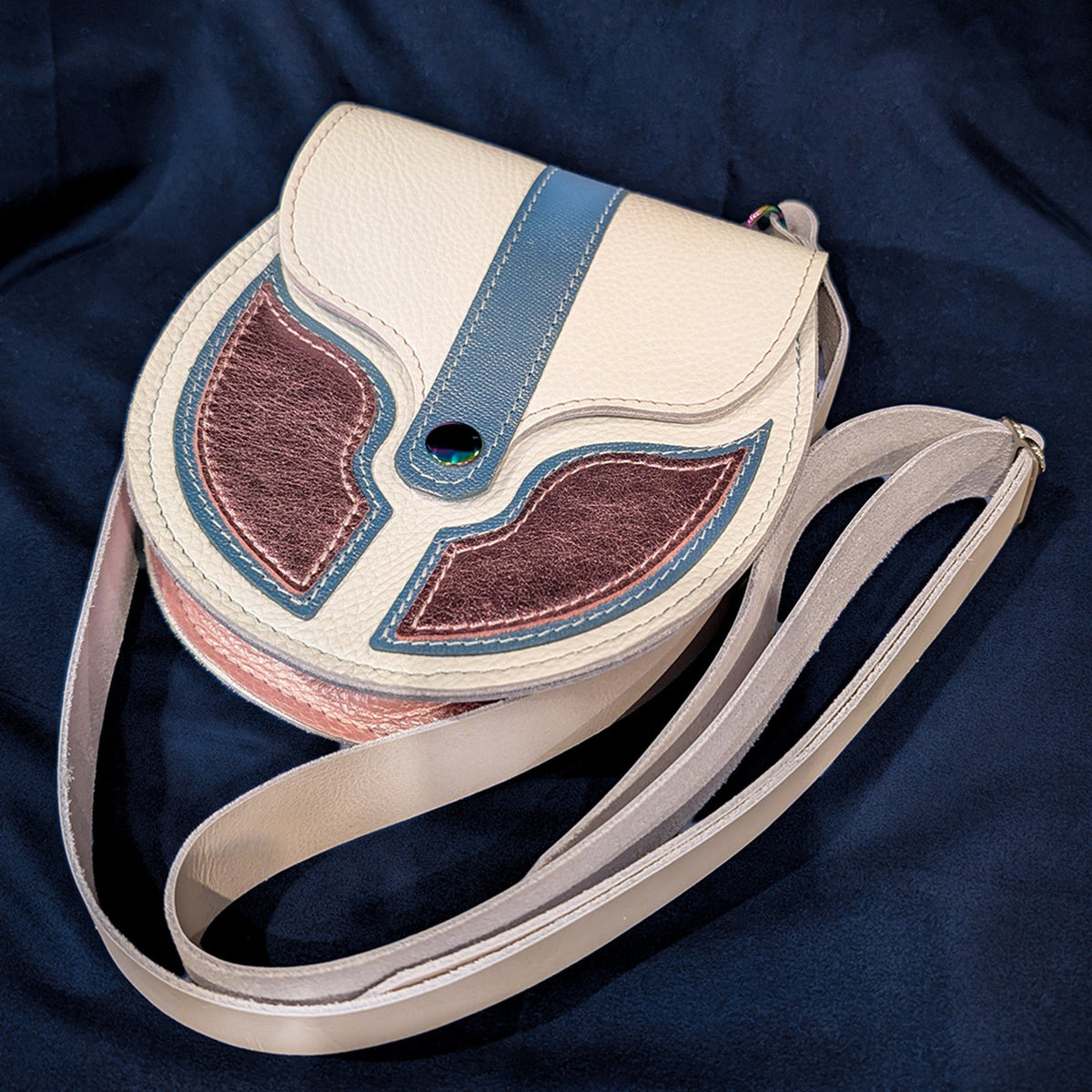 A white leather sporran bag with a blue strap detail and pink wing-shaped appliques, resting on a dark navy blue fabric background.