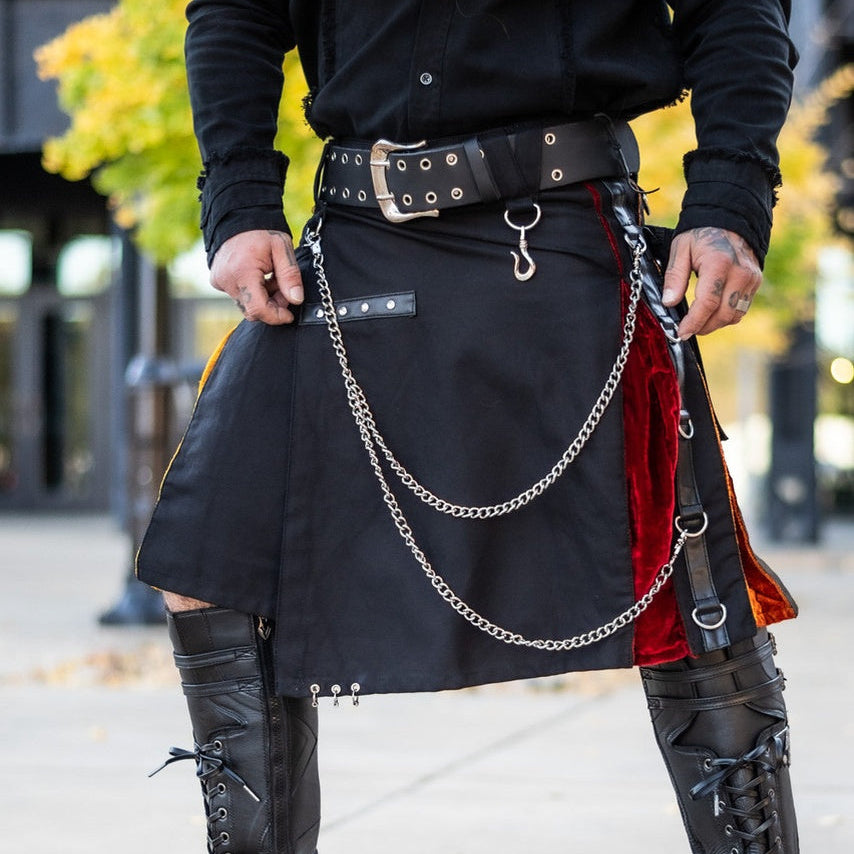 A person outdoors wearing a black gothic-style kilt with silver chain details, a black belt, a red fabric panel, and black lace-up knee-high boots.