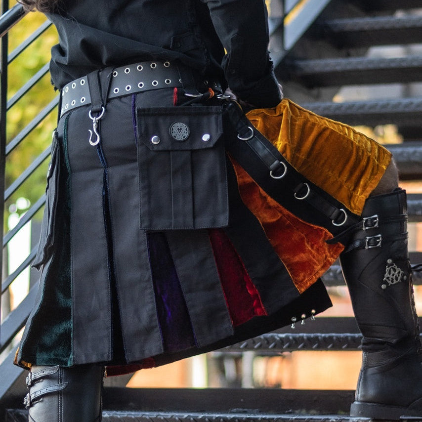 A person wearing a black gothic-style kilt with multiple utility pockets, silver studs, and colorful fabric panels, standing on outdoor metal stairs.