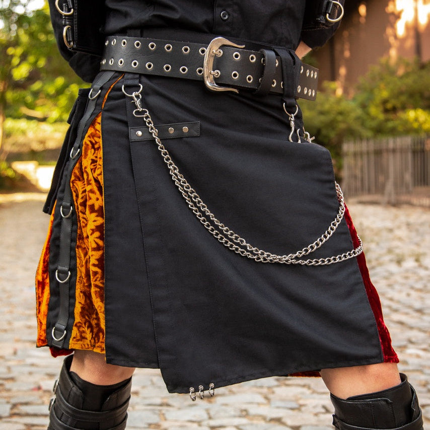 A person wearing a black asymmetrical kilt with orange patterned fabric panels, red side panels, a black studded belt, and silver chain details, standing outdoors on a cobblestone path.