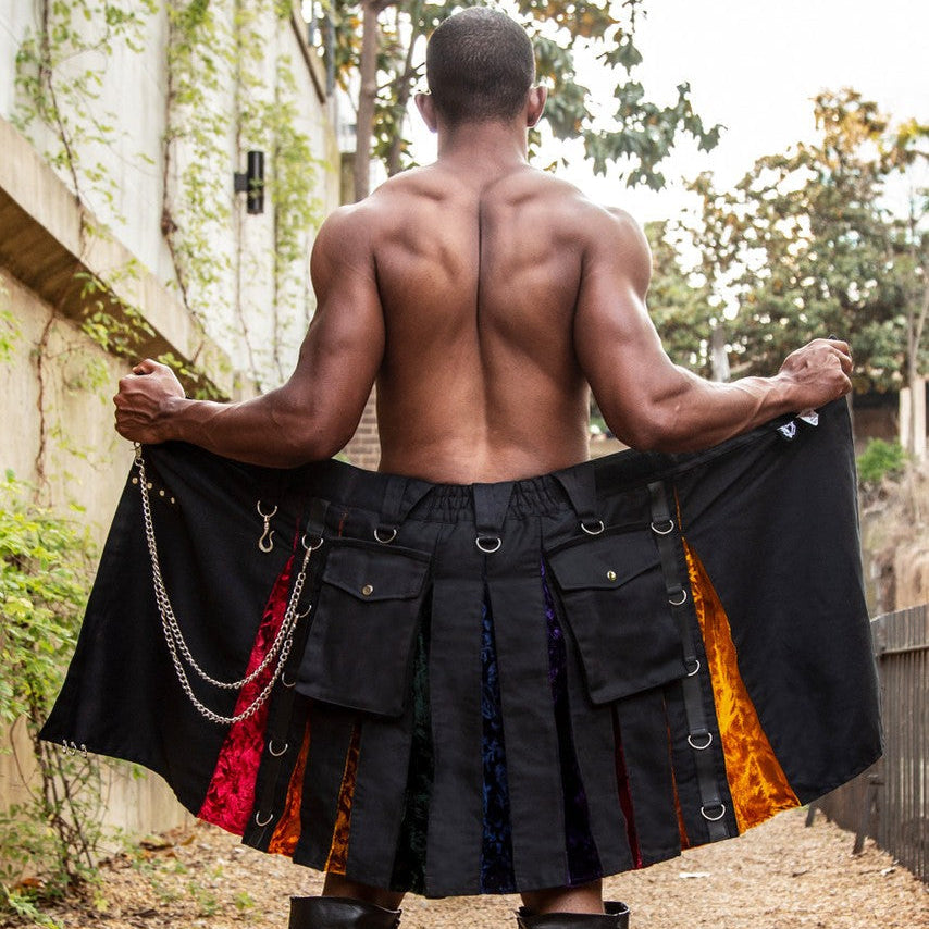 A shirtless man stands outdoors facing away from the camera, holding open a black pleated kilt-style garment with multiple flap pockets and colorful striped fabric panels in rainbow colors. 