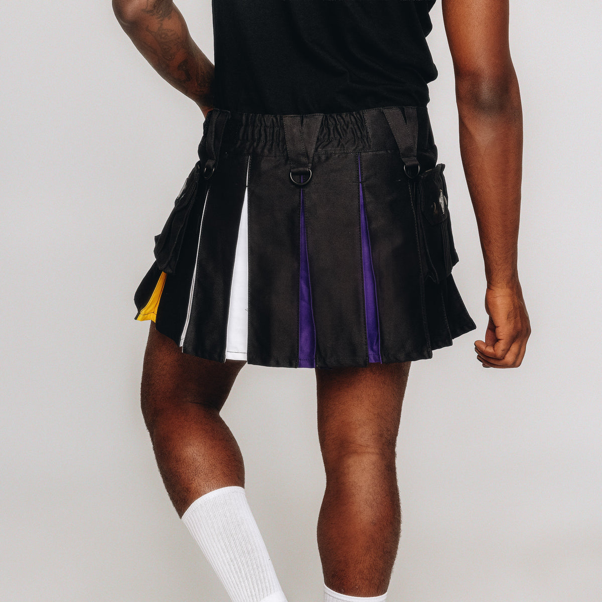 A model wearing a black pleated mini kilt with vertical panels in white, purple, and yellow, styled with a black sleeveless top and white knee-high socks against a light gray studio background.
