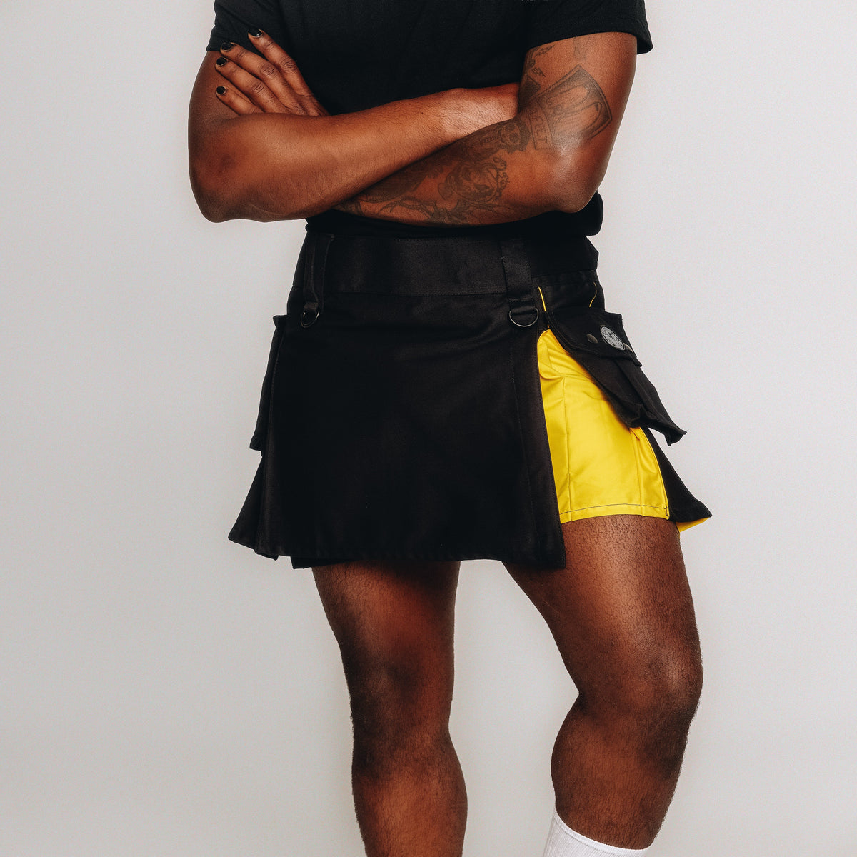 A model wearing a black layered kilt with a bright yellow side panel and black athletic shorts underneath, paired with a black t-shirt against a plain light gray background.
