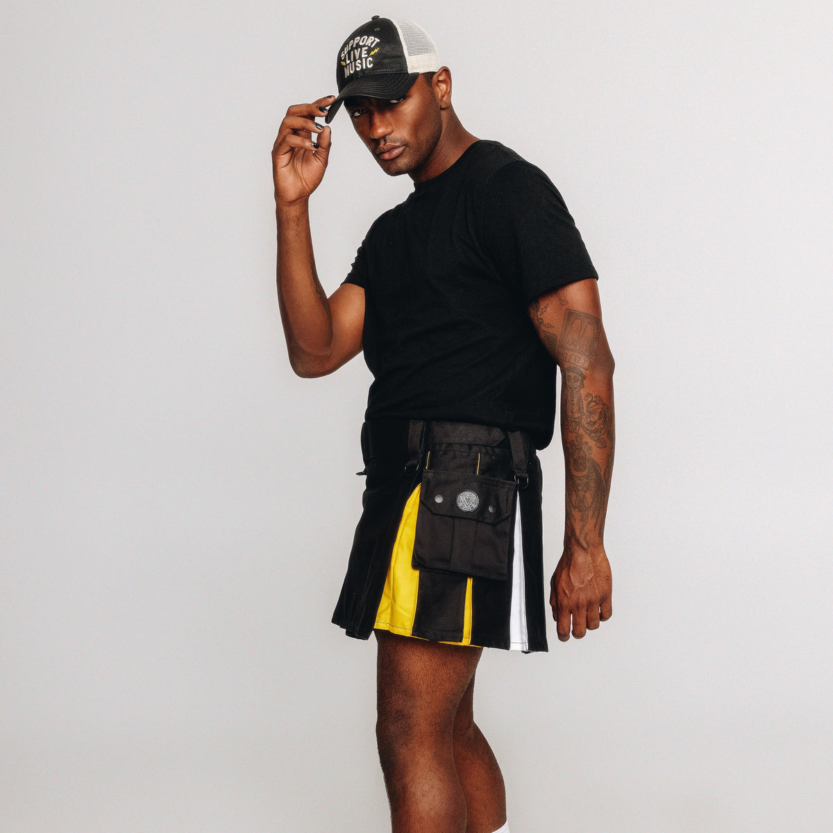 Person wearing a black t-shirt, black and nonbinary pride inspired kilt, white socks, and black shoes on a white background