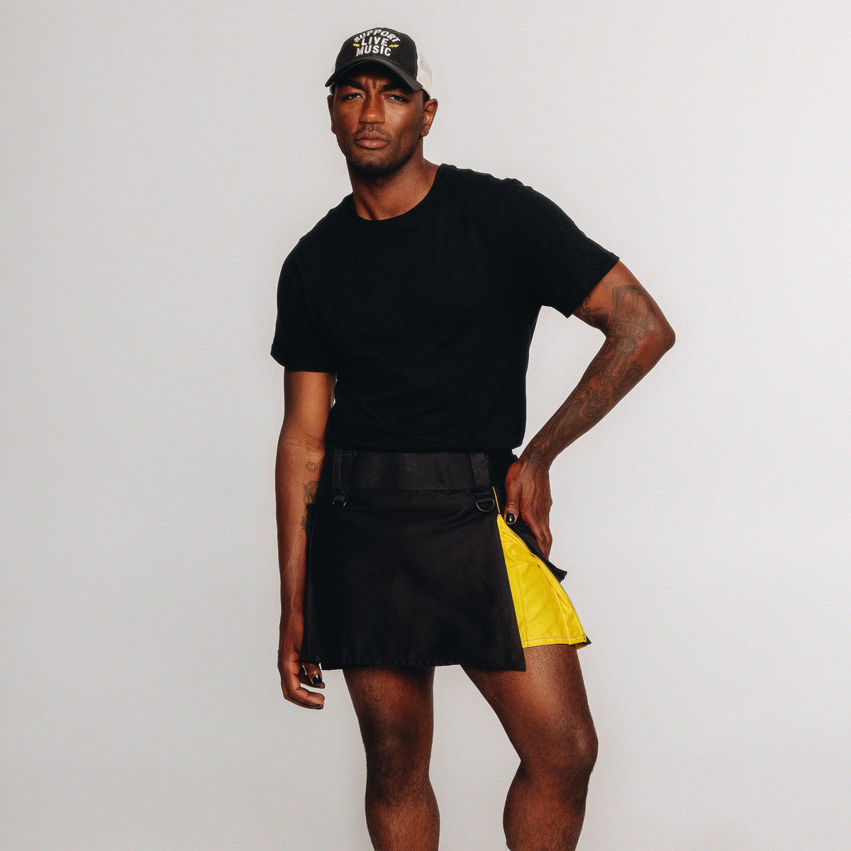 A person modeling a black kilt with a bright yellow side panel, and a black baseball cap with white text, standing against a plain light gray background.