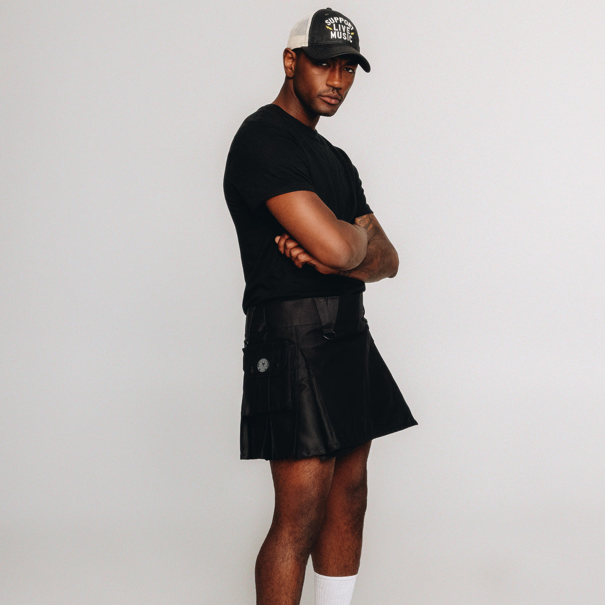 A man wearing a black t-shirt, a black pleated mini kilt, white socks, and a black and white trucker hat standing against a light gray studio background.