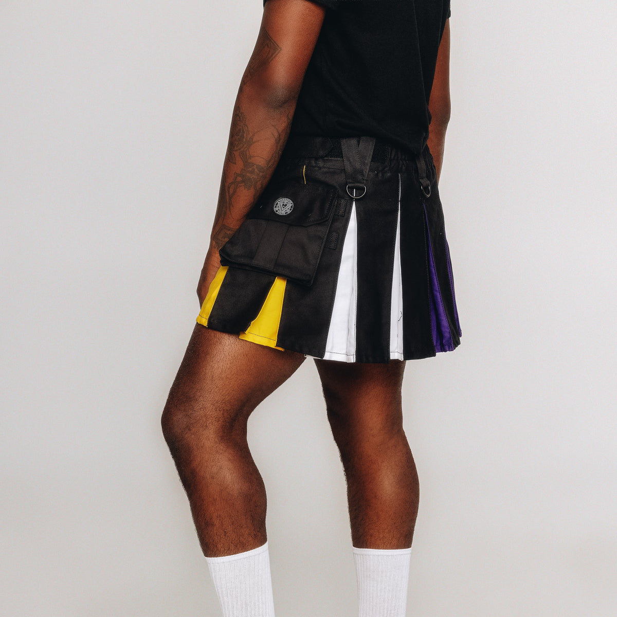 A model wearing a black pleated utility mini kilt with vertical panels in yellow, white, and purple, styled with a black t-shirt and white socks against a light gray background.