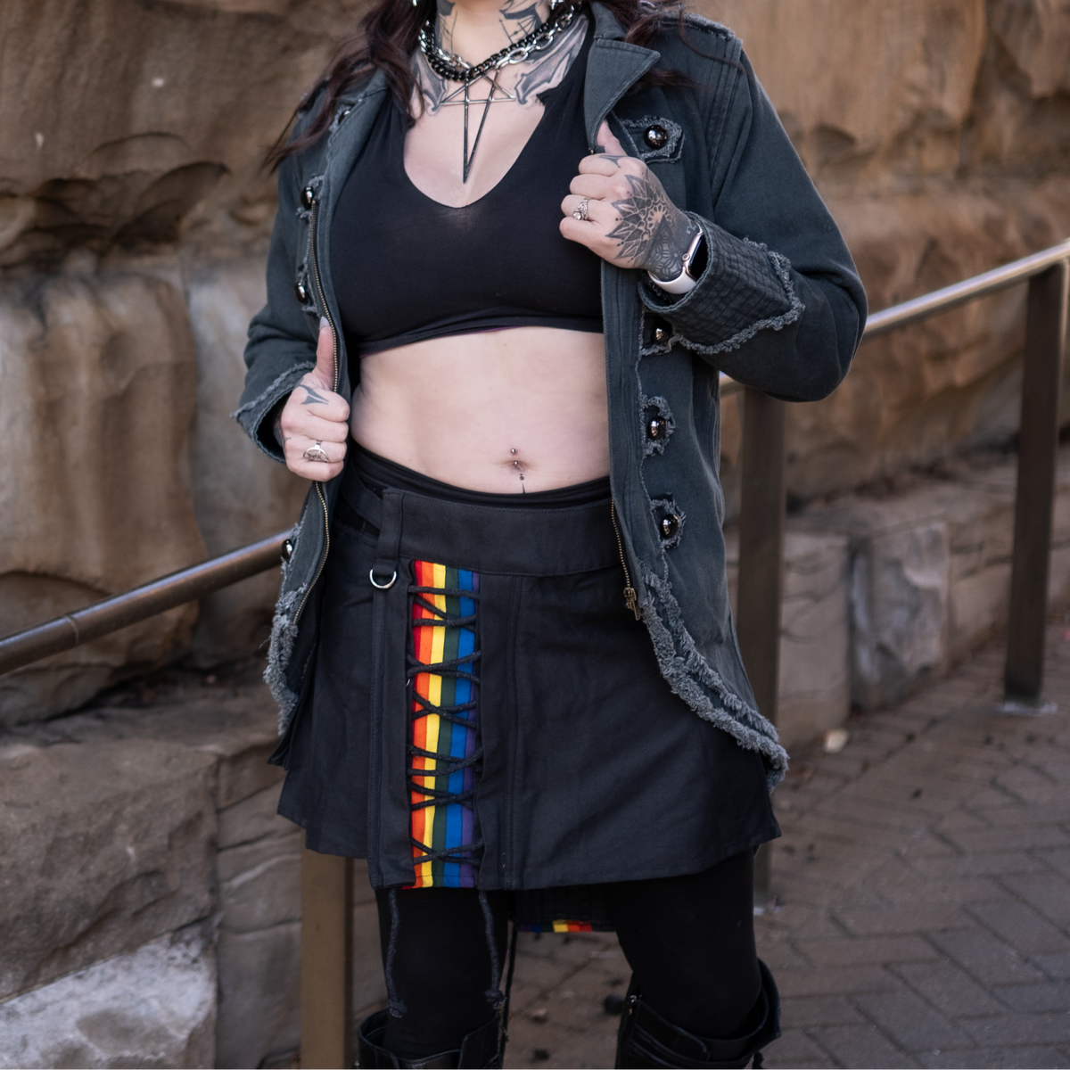 Woman in a black outfit with a black kilt with rainbow stripes standing against a stone wall.