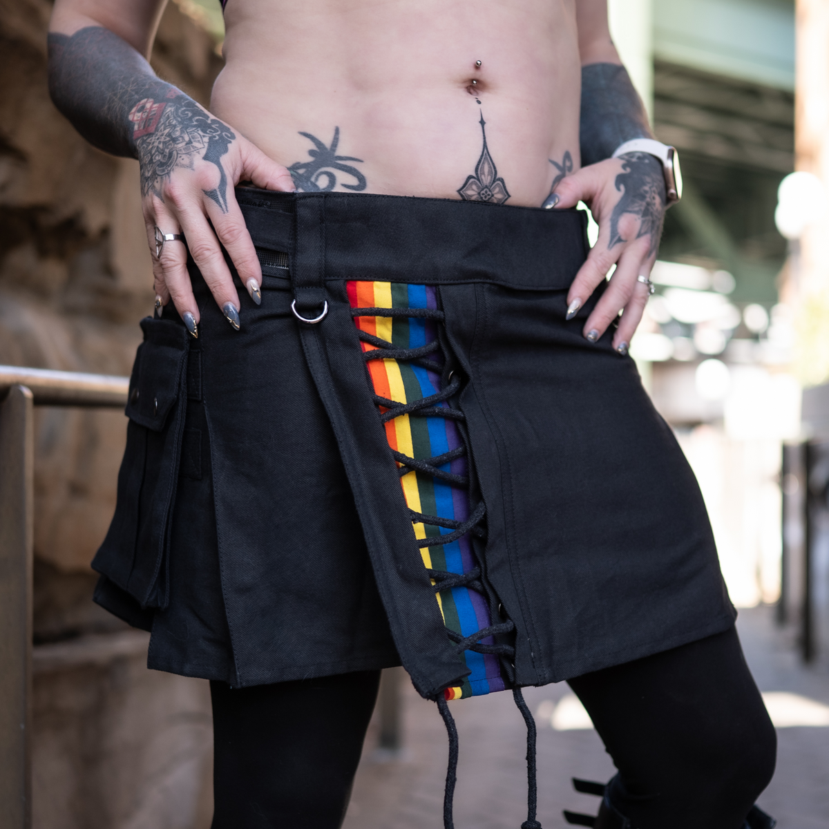 Person wearing a black skirt with lgbtq rainbow inset stripes, black laces and tattoos on arms and hands.
