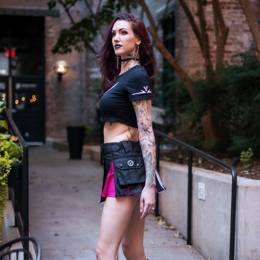 A woman stands on an outdoor sidewalk wearing a black cropped short-sleeve top, a black kilt with a bright pink inner layer, and tall black lace-up platform boots.
